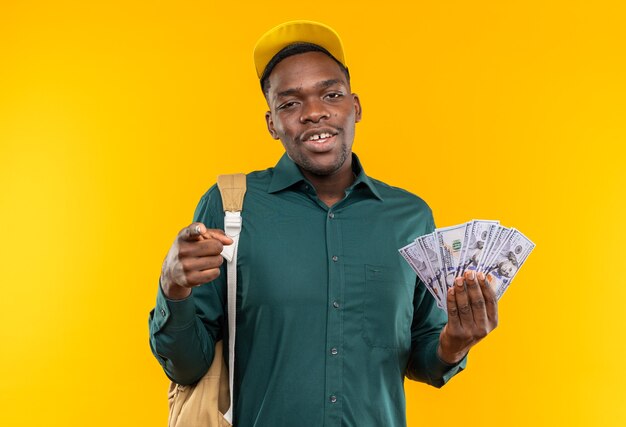 Pleased young afro-american student with cap and backpack holding money and pointing at front