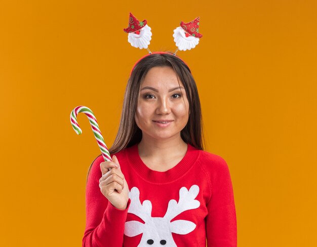 Pleased young asian girl wearing christmas hair hoop with sweater holding christmas candy isolated on orange background