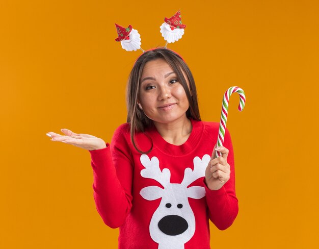 オレンジ色の背景で隔離の手を広げてクリスマスキャンディーを保持しているセーターとクリスマスの髪のフープを身に着けている若いアジアの女の子を喜ばせる