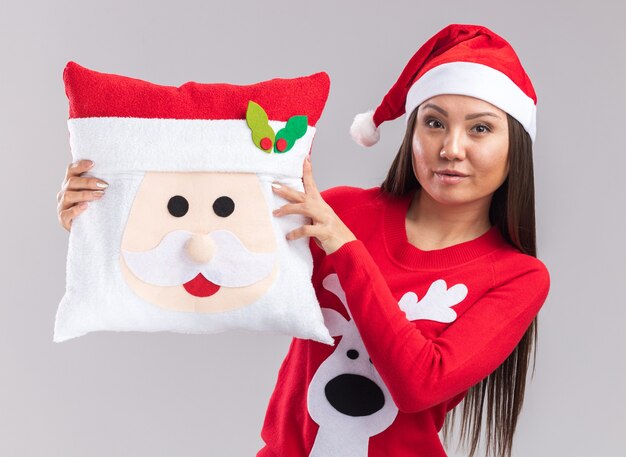 Довольная молодая азиатская девушка в новогодней шапке со свитером, держащая рождественскую подушку на белом фоне