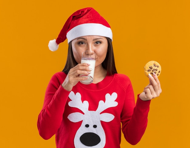 오렌지 배경에 고립 된 쿠키와 음료 우유를 들고 스웨터와 크리스마스 모자를 쓰고 기쁘게 젊은 아시아 여자