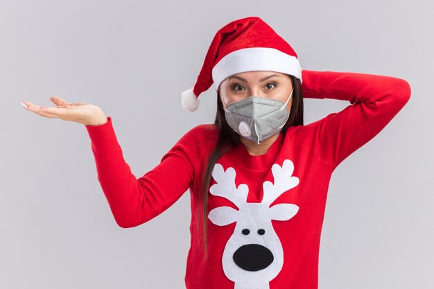 白い壁で隔離の頭に手を置くセーターと医療マスクとクリスマスの帽子をかぶって喜んで若いアジアの女の子