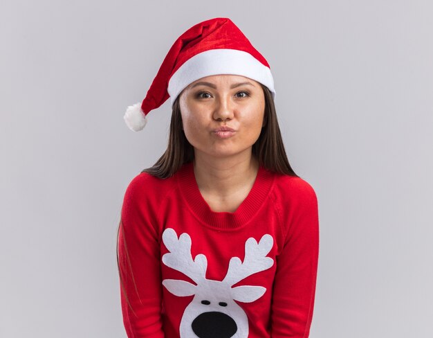 Довольная молодая азиатская девушка в новогодней шапке со свитером показывает жест поцелуя на белом фоне