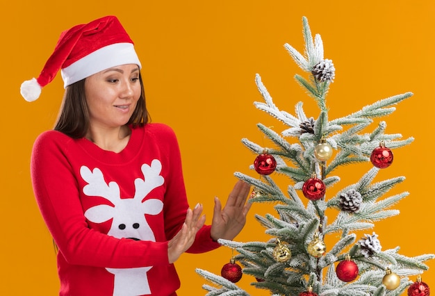 Pleased young asian girl wearing christmas hat with sweater standing nearby christmas tree isolated on orange background