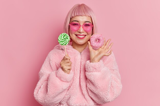 Pleased young Asian woman with pink bob hair smiles gently holds lollipop and delicious doughnut