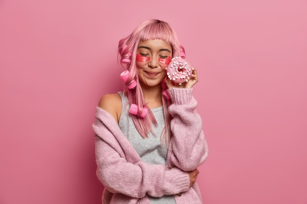 Pleased young Asian woman with pink hair, closes eyes holds delicious doughnuts near face, applies collagen patches under eyes, makes curly hairstyle