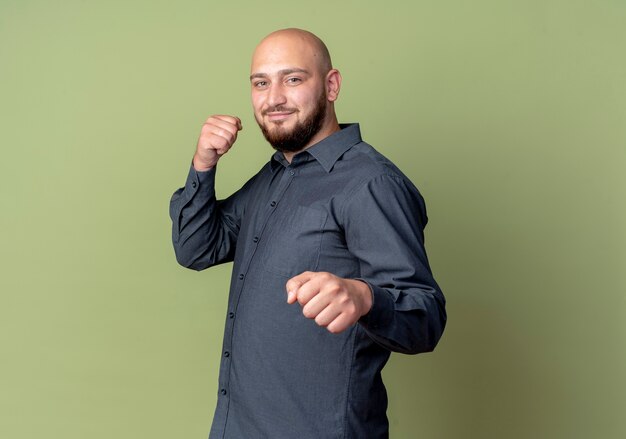 Довольный молодой лысый человек из колл-центра делает боксерский жест на камеру, изолированные на оливково-зеленом фоне с копией пространства
