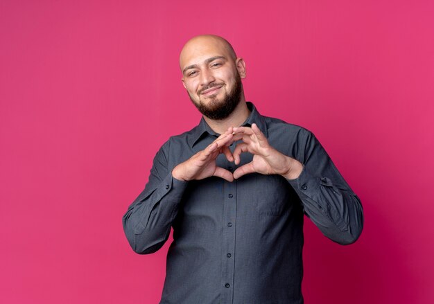 Pleased young bald call center man doing heart sign isolated on crimson background with copy space