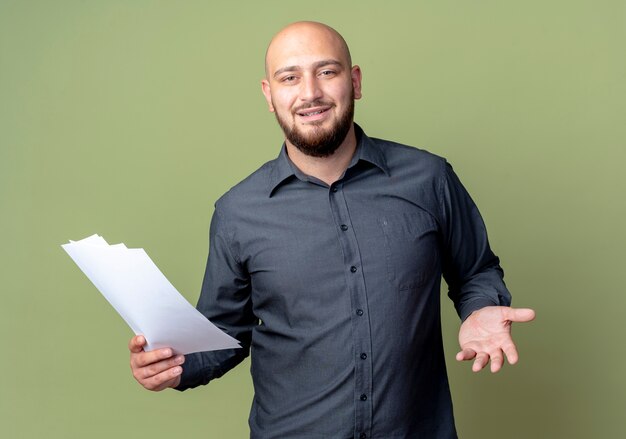Pleased young bald call center man holding documents and showing empty hand isolated on olive green background