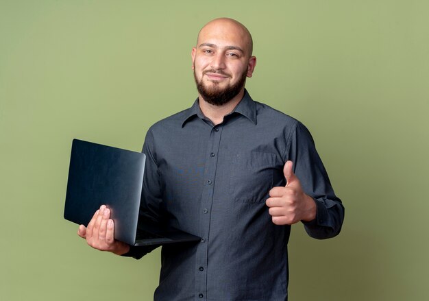Довольный молодой лысый человек колл-центра держит ноутбук и показывает большой палец вверх изолирован на оливково-зеленом фоне с копией пространства