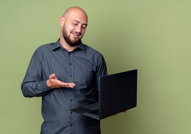Pleased young bald call center man holding looking at and pointing at laptop isolated on olive green background with copy space