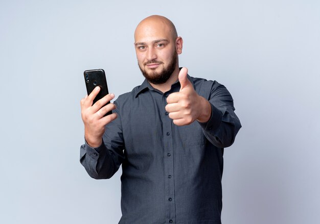 Pleased young bald call center man holding mobile phone and showing thumb up isolated on white background with copy space