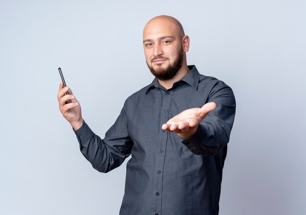 Pleased young bald call center man holding mobile phone and stretching out hand at camera isolated on white background with copy space