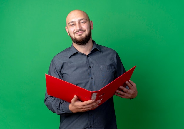 Pleased young bald call center man holding open folder isolated on green background with copy space