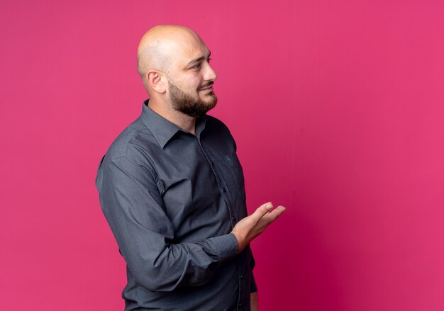 Pleased young bald call center man looking straight and pointing with hand at side isolated on crimson background with copy space