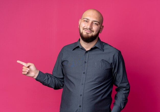 Pleased young bald call center man pointing at side isolated on crimson background
