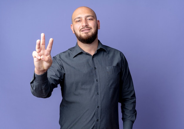 Pleased young bald call center man showing three with hand isolated on purple background with copy space
