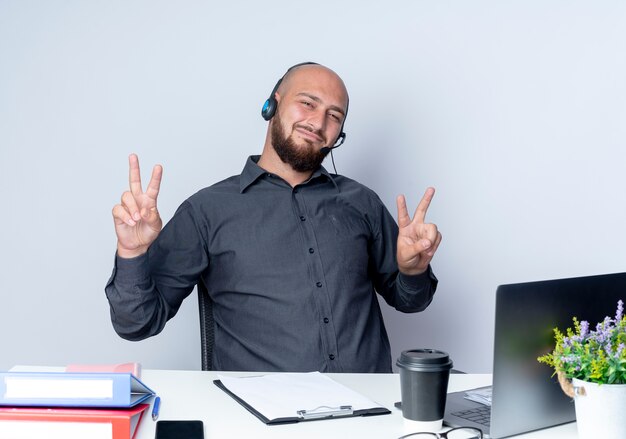Pleased young bald call center man wearing headset sitting at desk with work tools doing peace signs isolated on white background