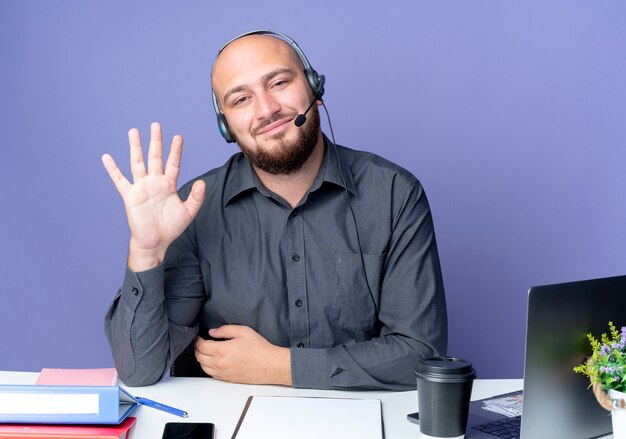 Pleased young bald call center man wearing headset sitting at desk with work tools gesturing hi at camera isolated on purple background