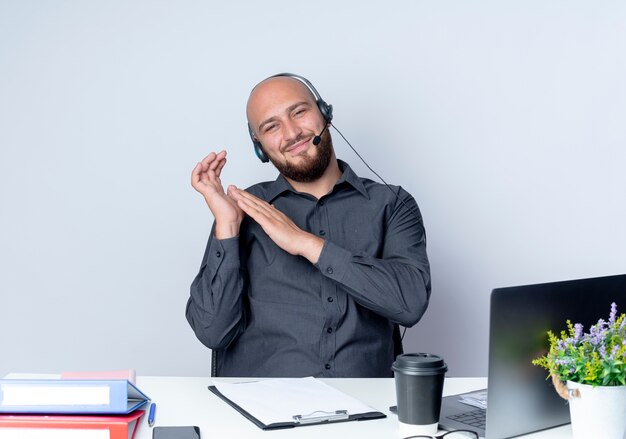 Free photo pleased young bald call center man wearing headset sitting at desk with work tools keeping hand near another one isolated on white background