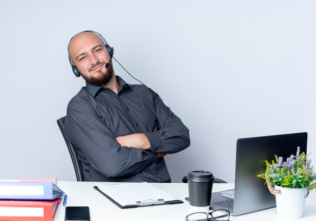 Pleased young bald call center man wearing headset sitting with closed posture at desk with work tools isolated on white background