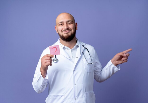 Pleased young bald male doctor wearing medical robe and stethoscope holding paper question mark and points at side