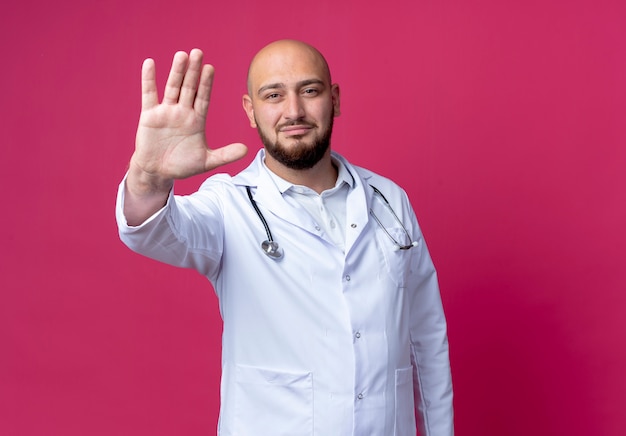 Pleased young bald male doctor wearing medical robe and stethoscope showing stop gesture isolated on pink wall