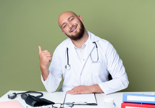 Pleased young bald male doctor wearing medical robe and stethoscope sitting at desk work with medical tools points at side isolated on green background