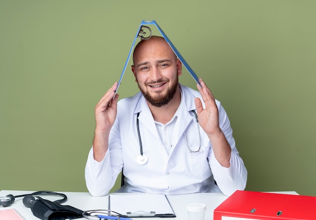 Foto gratuita lieto giovane medico maschio calvo che indossa una tunica medica e uno stetoscopio seduto alla scrivania