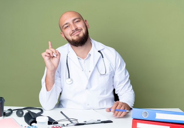 Довольный молодой лысый мужчина-врач в медицинском халате и стетоскопе сидит за столом