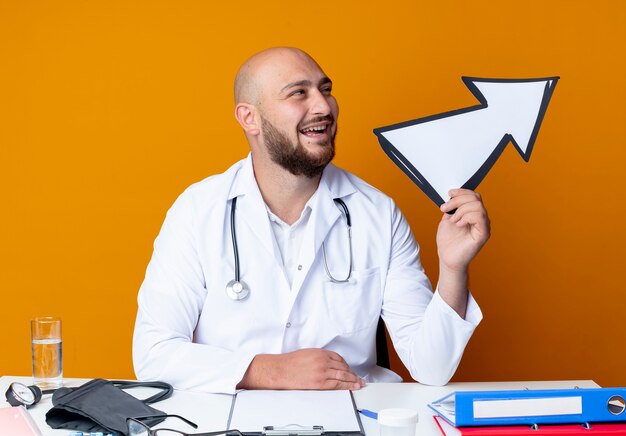 Довольный молодой лысый врач-мужчина в медицинском халате и стетоскопе сидит за рабочим столом с медицинскими инструментами, держа знак направления, изолированный на оранжевом фоне