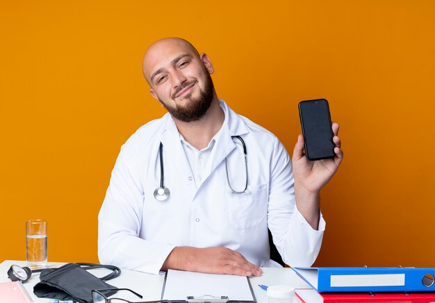 Довольный молодой лысый мужчина-врач в медицинском халате и стетоскопе сидит за рабочим столом с медицинскими инструментами, держа телефон, изолированный на оранжевом фоне