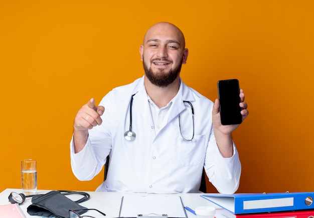 Felice giovane maschio calvo medico indossando abito medico e stetoscopio seduto alla scrivania con strumenti medici tenendo il telefono e mostrando il gesto isolato su sfondo arancione