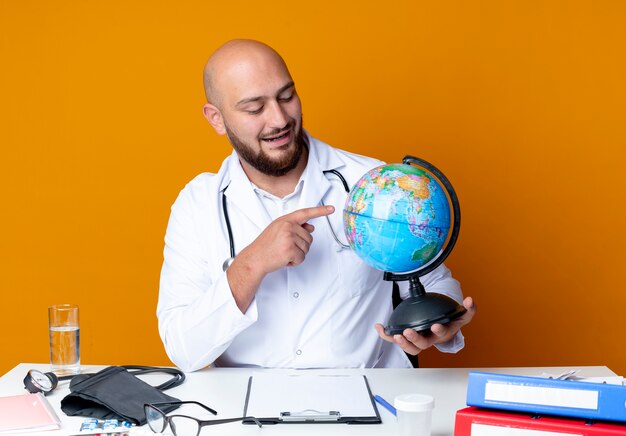 Pleased young bald male doctor wearing medical robe and stethoscope sitting at work desk with medical tools holding and points at globe isolated on orange background