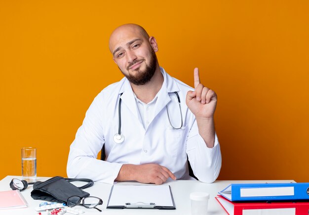 Pleased young bald male doctor wearing medical robe and stethoscope sitting at work desk with medical tools points at up isolated on orange background