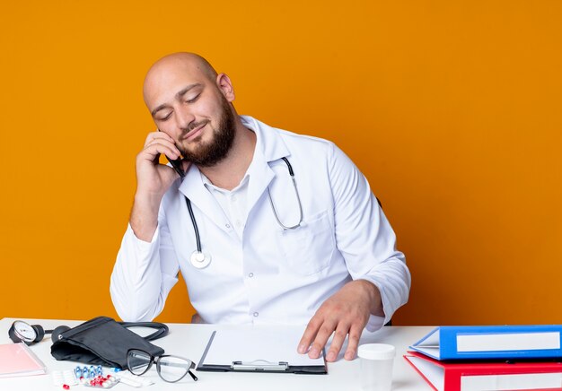 Pleased young bald male doctor wearing medical robe and stethoscope sitting at work desk with medical tools speaks on phone isolated on orange background