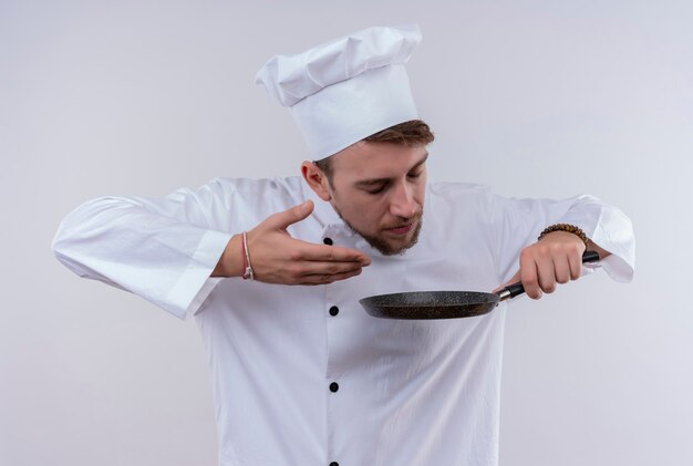 A pleased young bearded chef man wearing white cooker uniform and hat enjoying the cooking smell while holding frying pan on a white wall