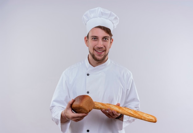 Un felice giovane chef barbuto uomo bianco che indossa un fornello uniforme e hat holding baguette con pane pagnotta mentre si guarda su un muro bianco