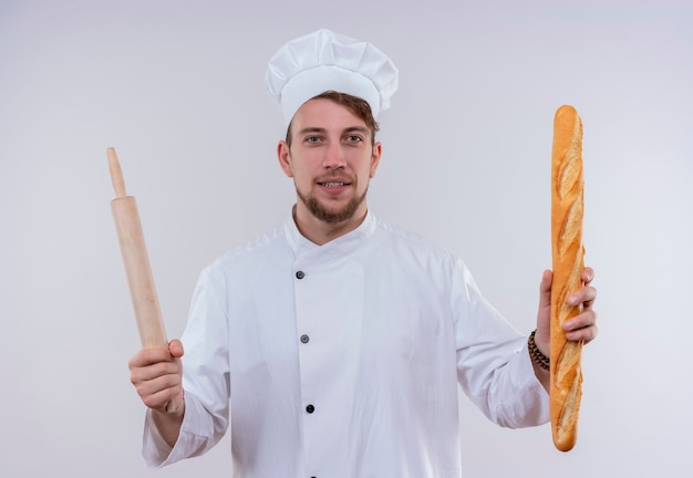 Un felice giovane chef barbuto uomo che indossa l'uniforme bianca del fornello e cappello che tiene il pane baguette con il mattarello mentre guarda su un muro bianco Foto Gratuite