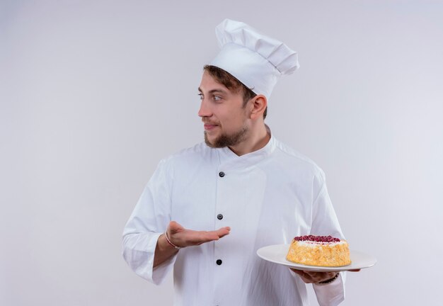 A pleased young bearded chef man wearing white cooker uniform and hat holding a plate with cake while looking side on a white wall