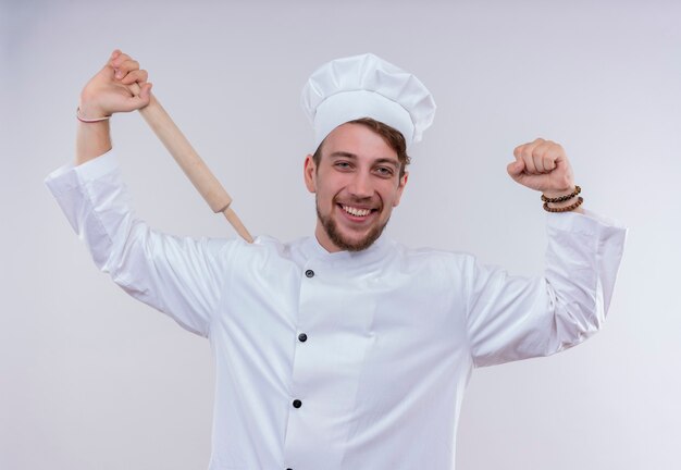 Довольный молодой бородатый шеф-повар в белой униформе и шляпе, держащий скалку в кулаке, глядя на белую стену
