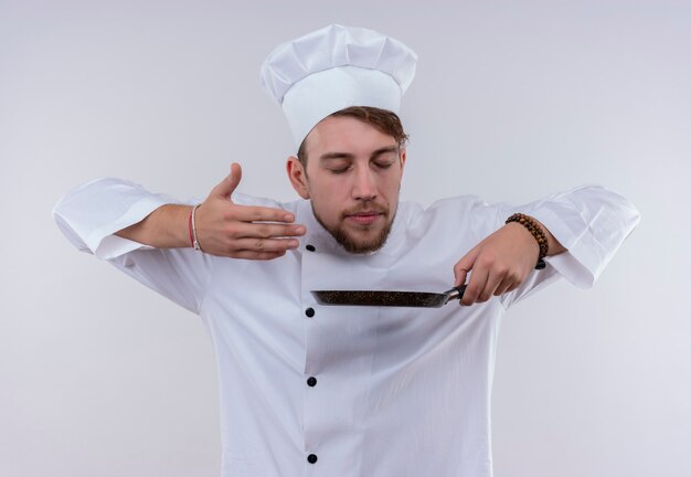 A pleased young bearded chef man wearing white cooker uniform and hat smelling frying pan on a white wall