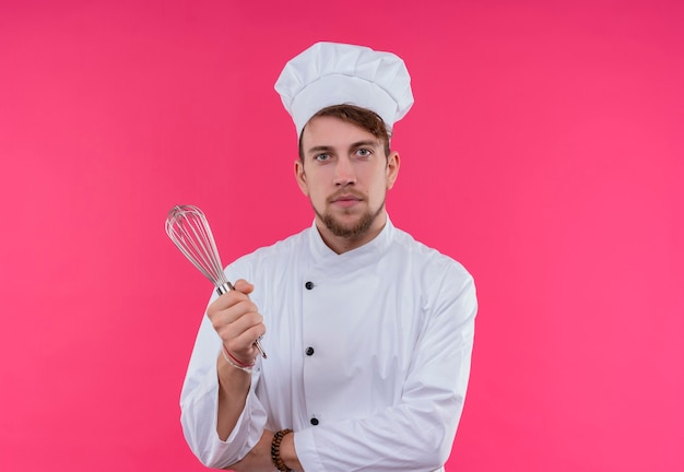 A pleased young bearded chef man in white uniform holding mixer spoon while looking on a pink wall