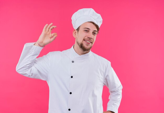 A pleased young bearded chef man in white uniform showing ok gesture on a pink wall