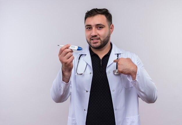 Pleased young bearded male doctor wearing white coat with stethoscope holding digital thermometer  smiling confident pointing to himself 