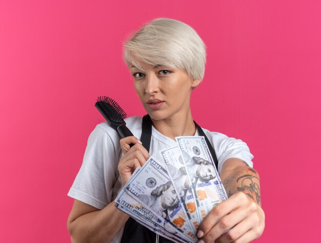 Pleased young beautiful female barber in uniform holding out comb with cash  isolated on pink wall