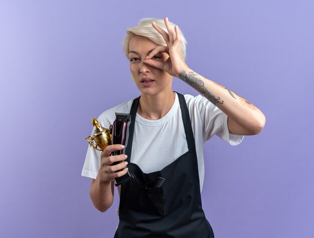 Pleased young beautiful female barber in uniform holding winner cup with hair clippers showing look gesture isolated on blue wall