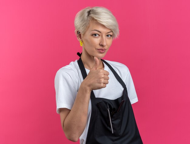 Pleased young beautiful female barber in uniform showing thumb up isolated on pink wall