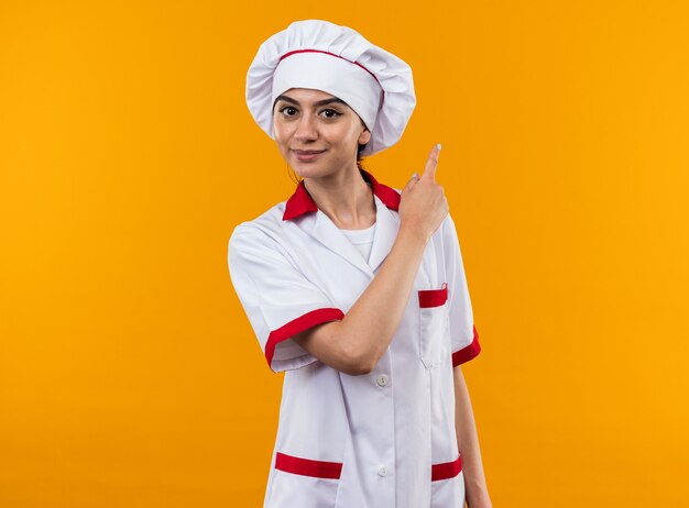 Pleased young beautiful girl in chef uniform points at behind 