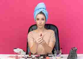 Бесплатное фото Довольная молодая красивая девушка сидит за столом с инструментами для макияжа, завернув волосы в полотенце, протягивая помаду, изолированную на розовой стене